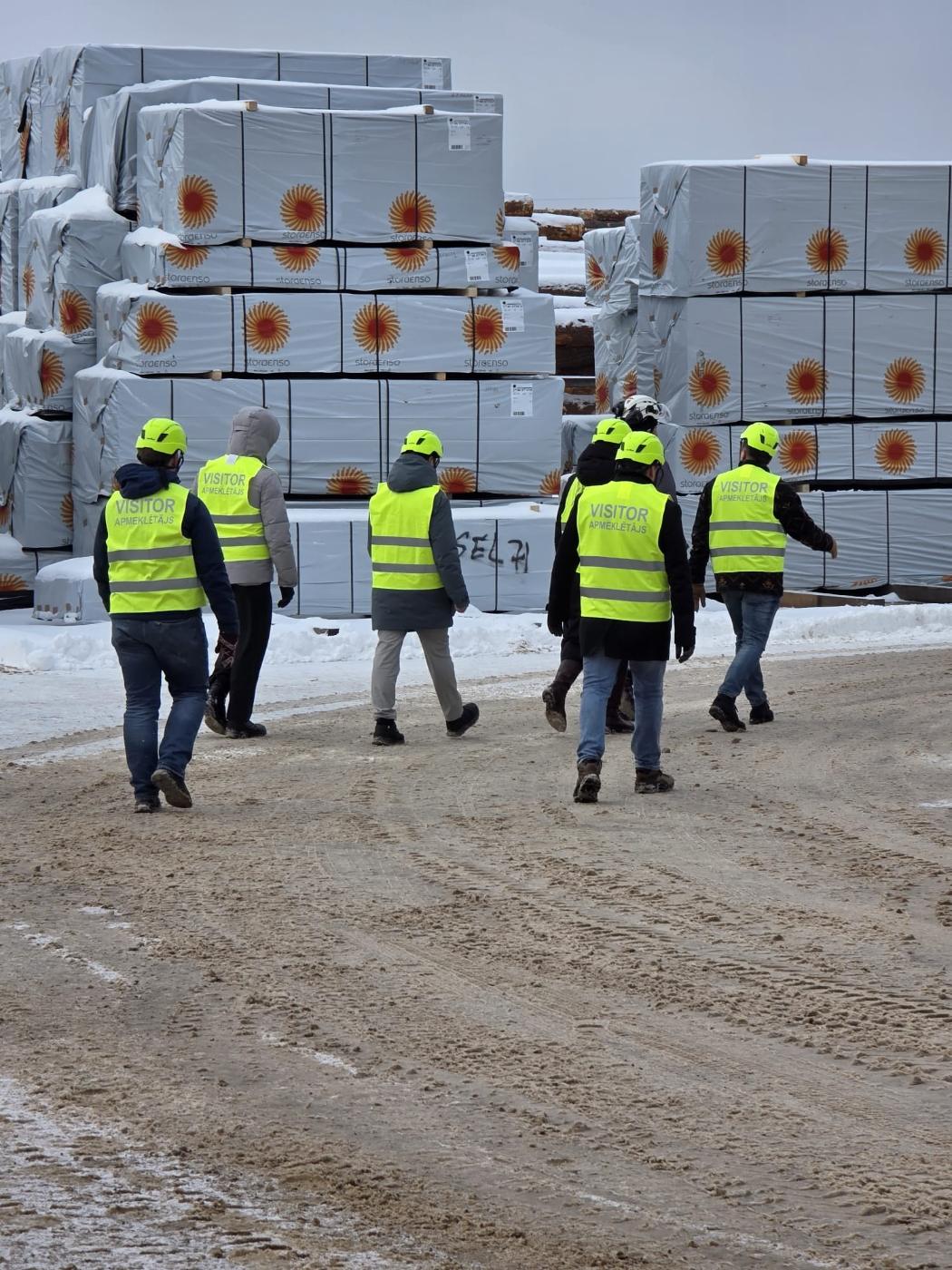 A group of visitors in hi-vis vests and helmets walking past wrapped packs of Stora Enso sawn timber in a snowy yard